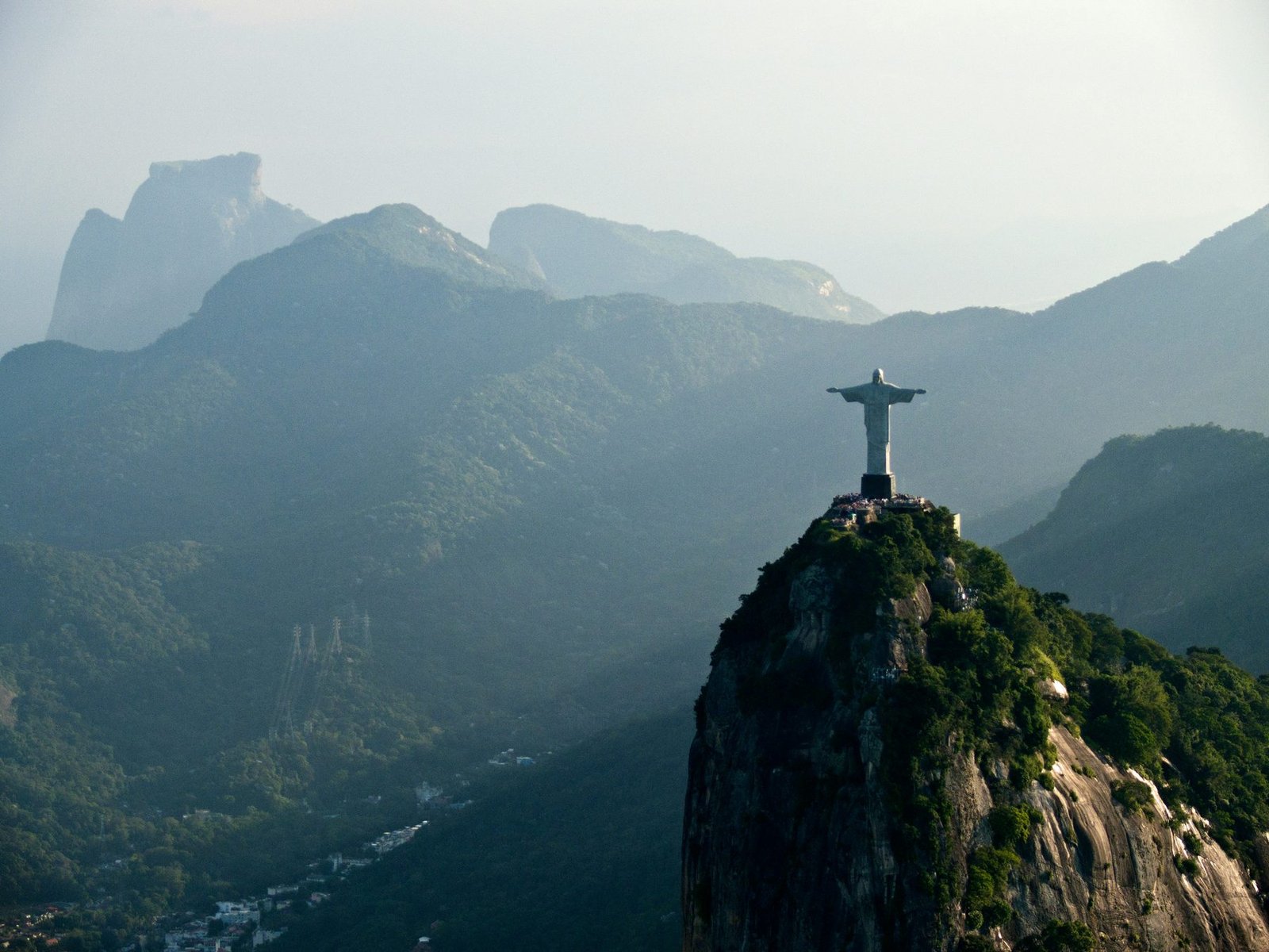A statue with outstretched arms stands on a rocky forested summit, with layers of hazy mountains behind