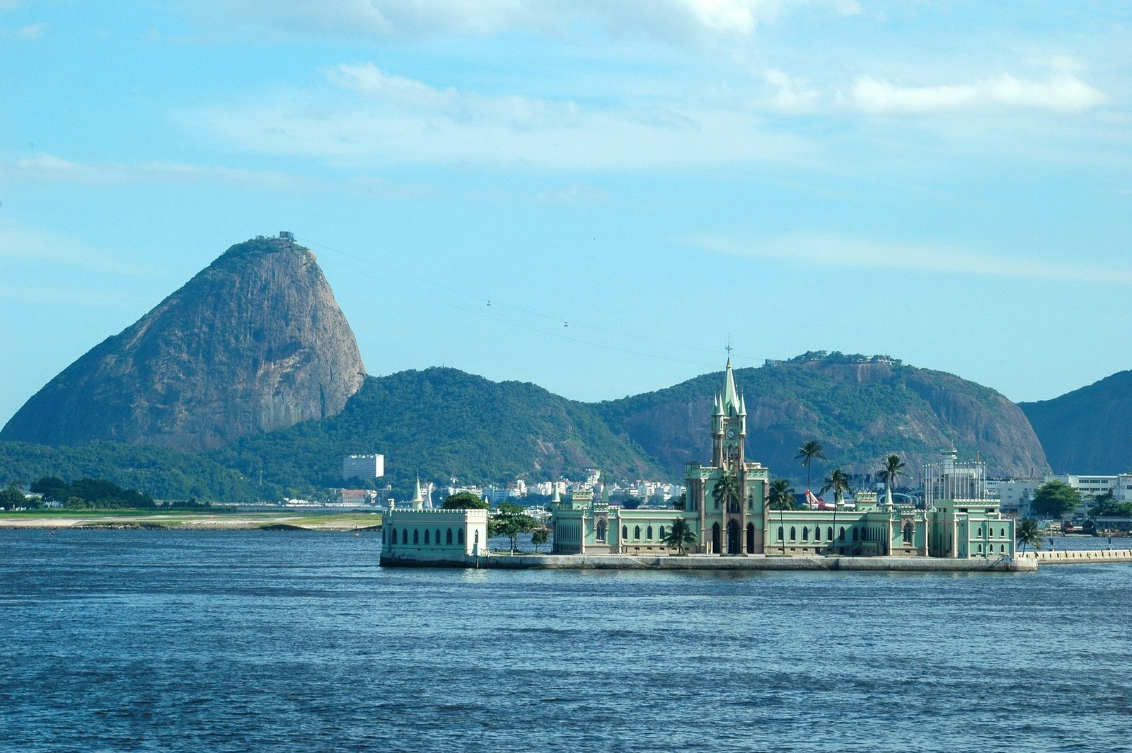 A green palace with a pointed central tower sits on a small island in a bay, below a tall rock peak and hills