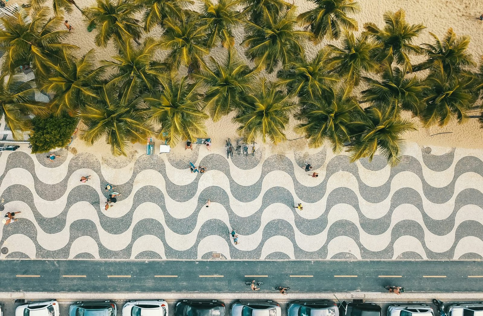 Overhead view of a black-and-white wave-patterned mosaic sidewalk between a row of palm trees on the sand and a line of parked cars