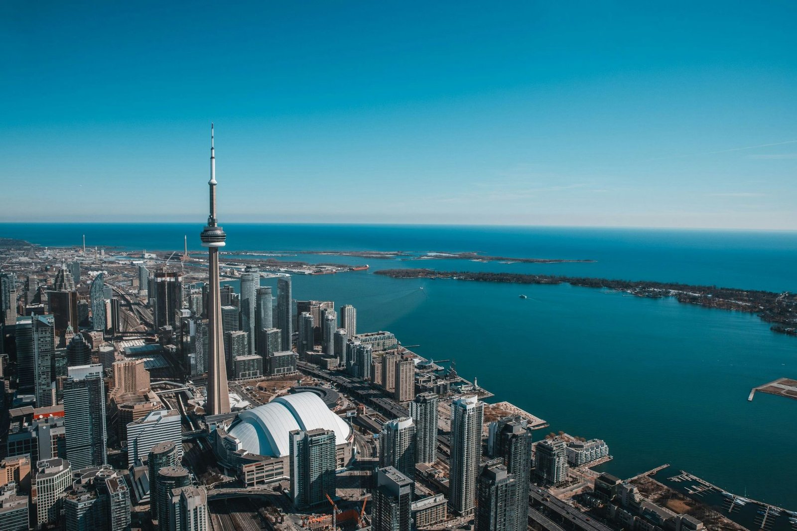 A tall concrete tower rises beside a white domed stadium above a turquoise lake edged by a long curving island