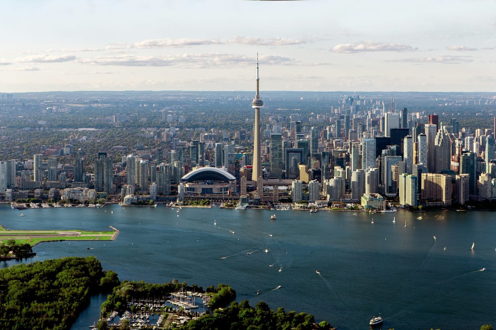 A needle-like tower and a white domed stadium stand among towers on a harbor dotted with boats, with wooded islands in the foreground