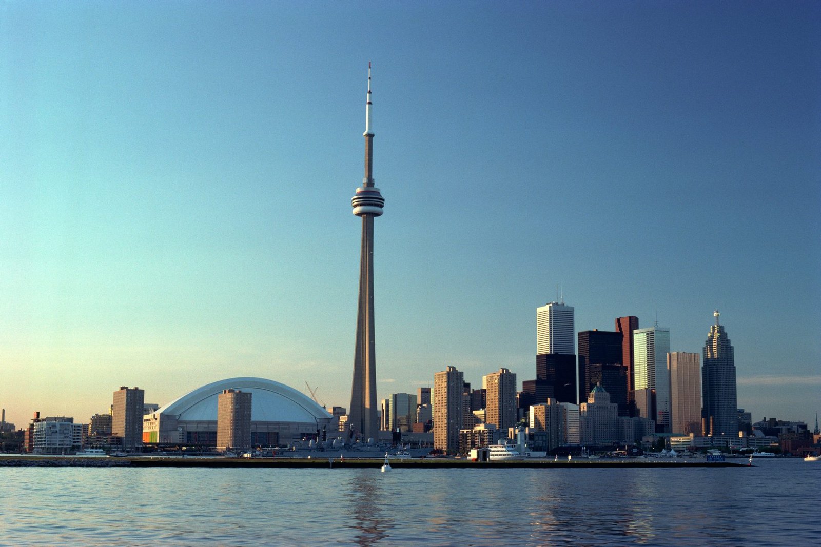 A tall concrete tower and a white dome stand beside a group of office towers above calm water in evening light
