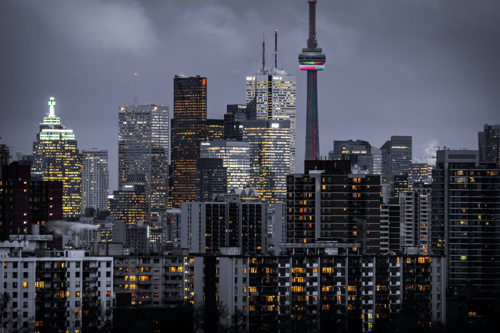 Lit office towers and a tall tower with a red-lit top rise behind dark apartment blocks under a purple night sky