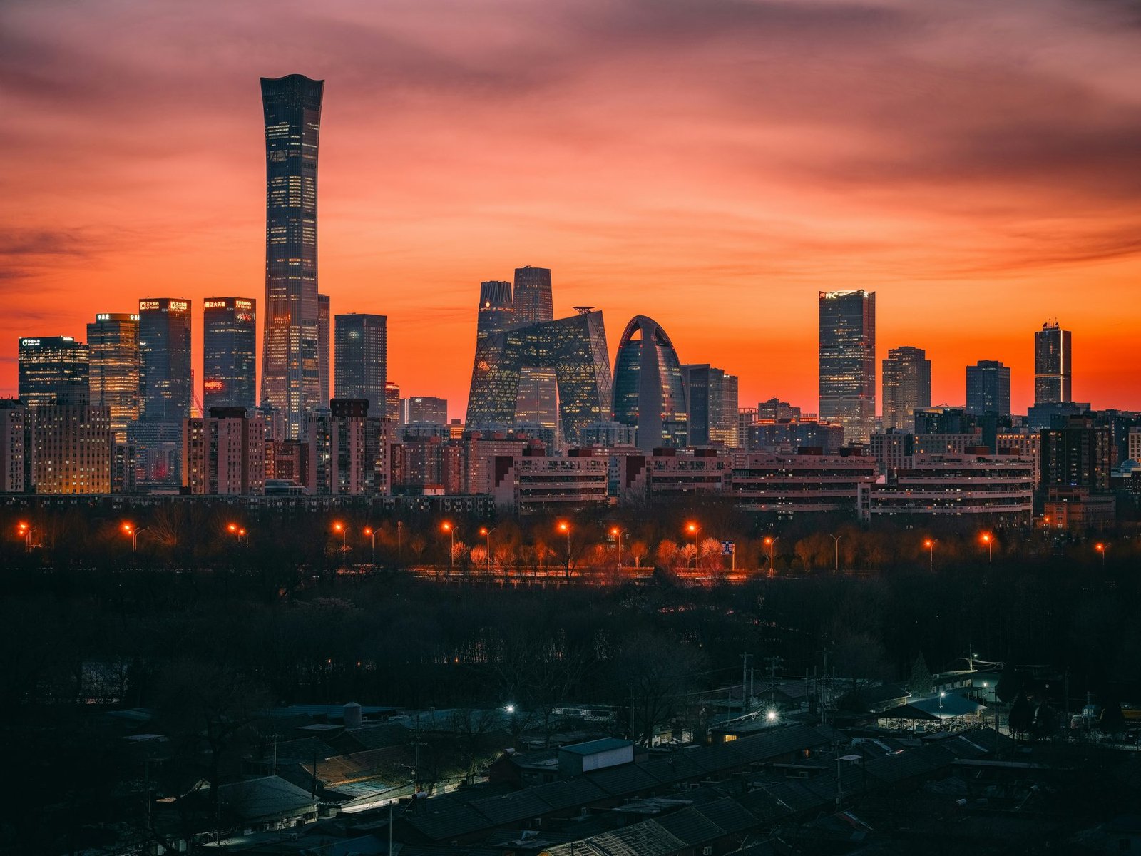 Dark skyscrapers, one far taller than the rest, stand against a red and orange sky above a dark foreground with scattered lights