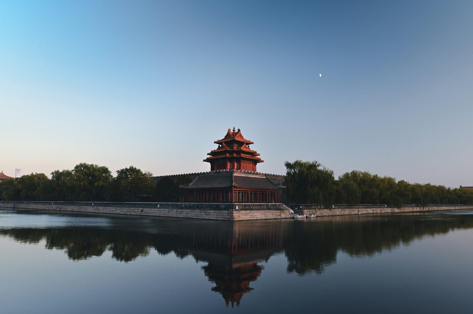A red-walled tower with layered roofs is mirrored in a still moat under a clear sky