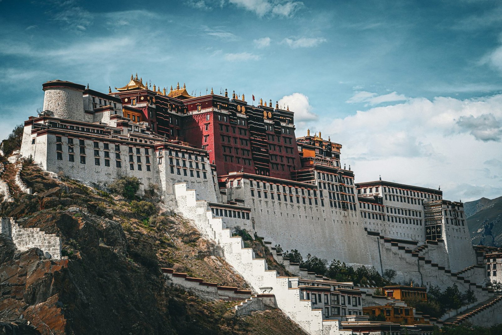 A white and red palace climbs a rocky hill with zigzag stairways under a cloudy sky