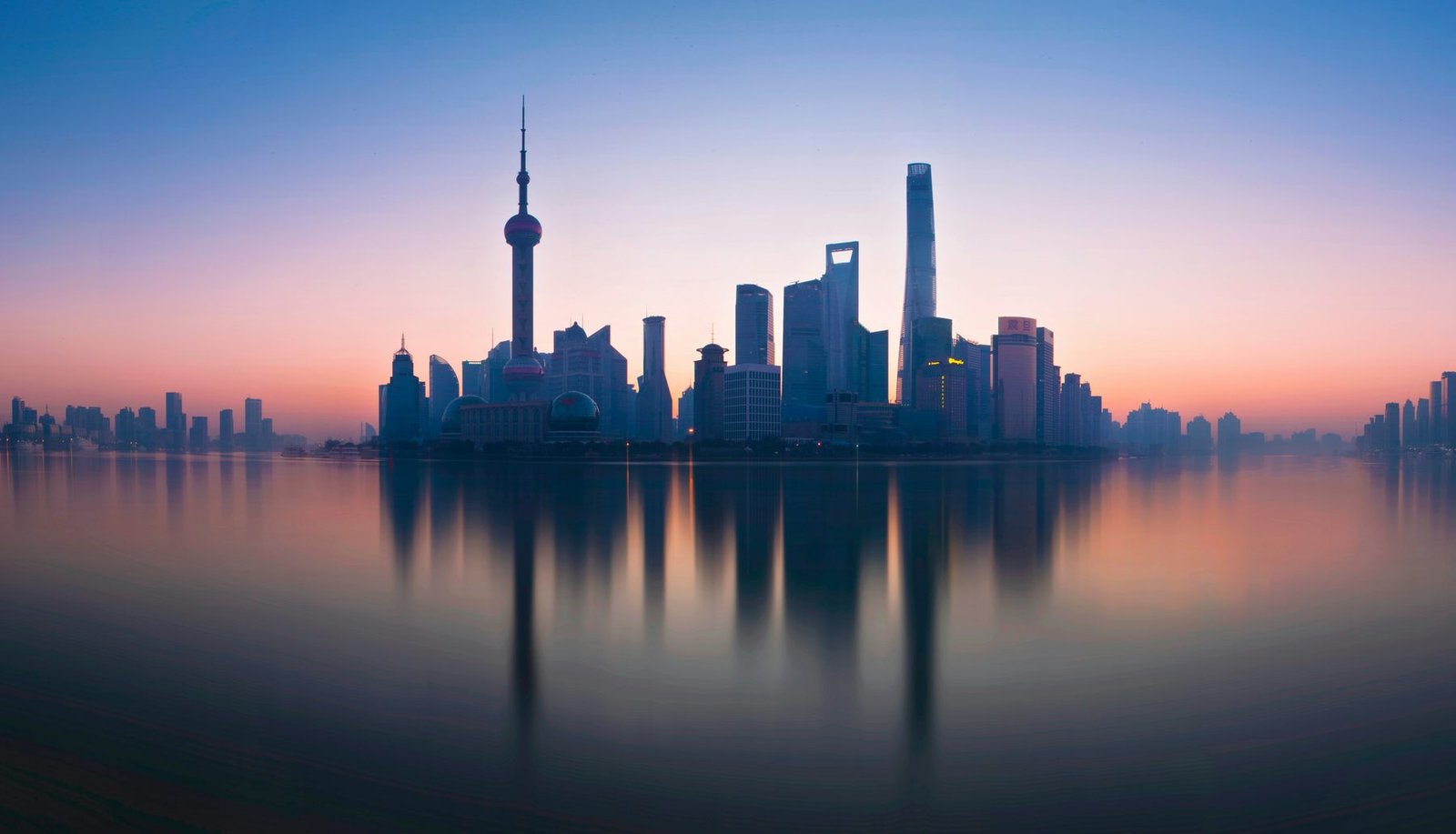 A skyline in silhouette is reflected in smooth water under a pink and blue sky