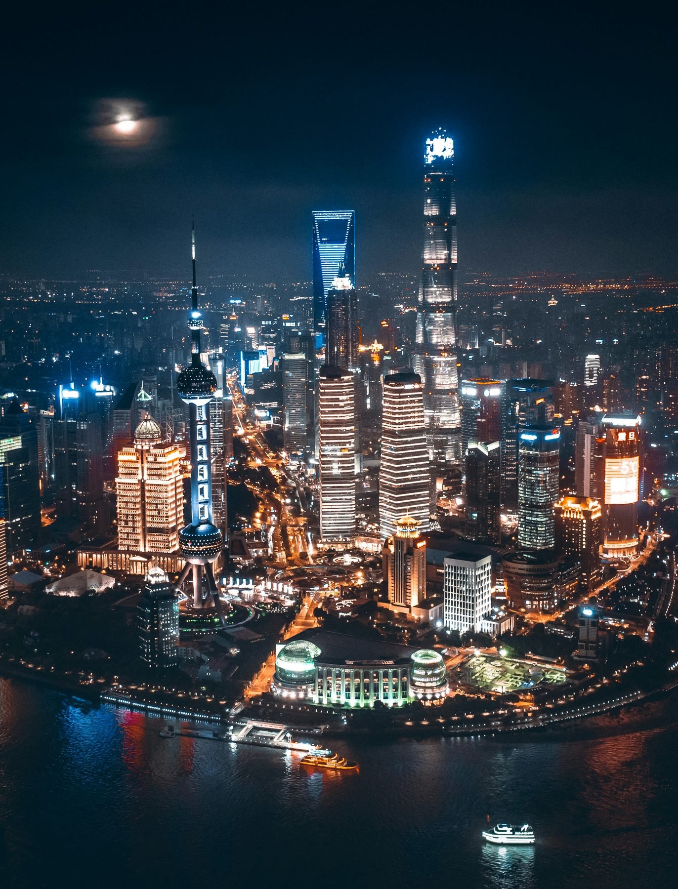 Lit skyscrapers and a tower with spheres rise from a riverside district beneath a small moon