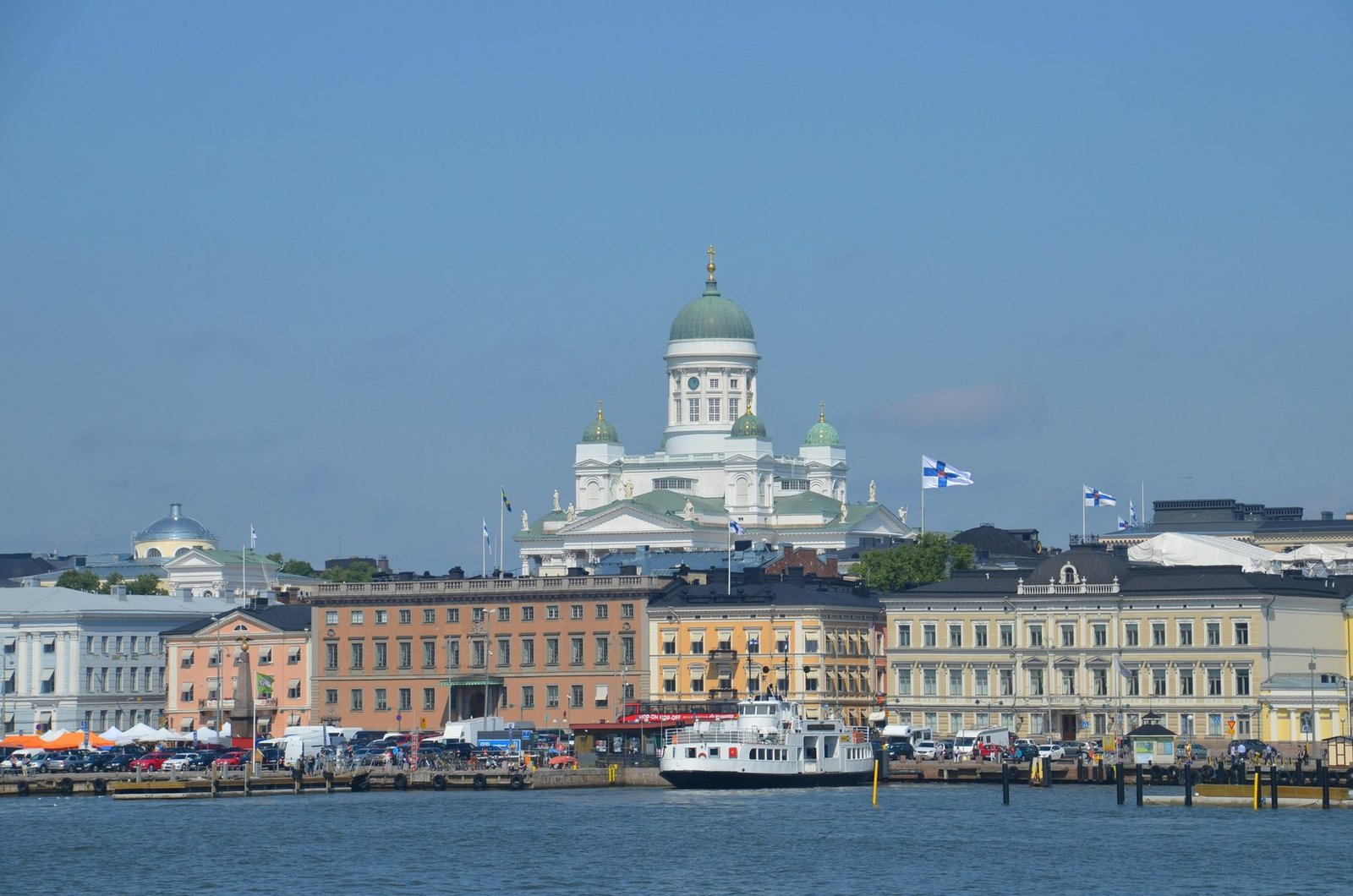 A white cathedral with a green dome rises above a row of pastel waterfront buildings, with a white ferry at the quay