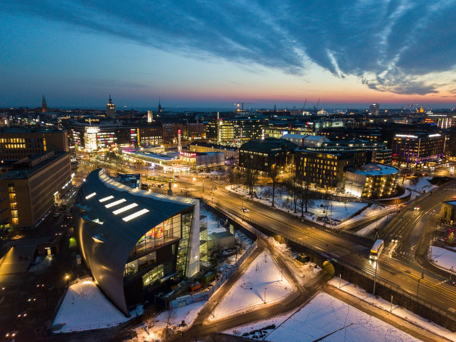 Lit streets with light trails curve around a dark building with a curved roof, among snow-covered ground and city blocks under a sunset sky
