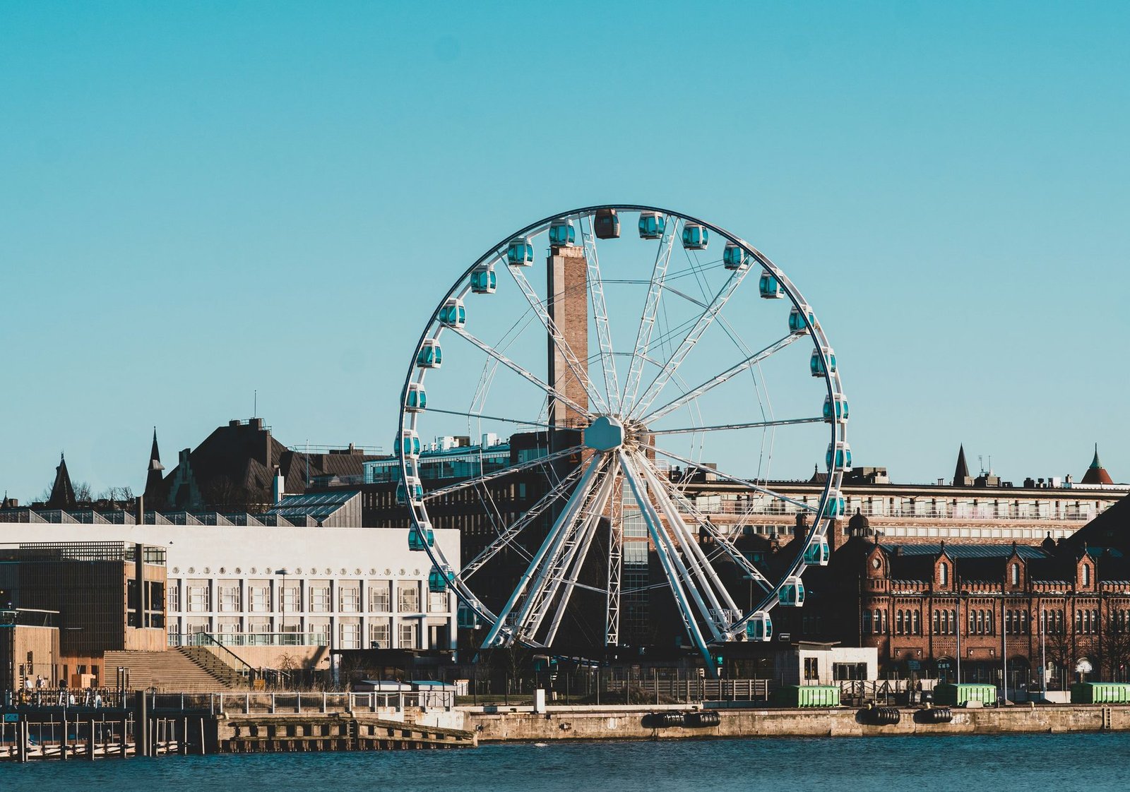 A large Ferris wheel with blue cabins stands on a quay beside low buildings under a clear sky