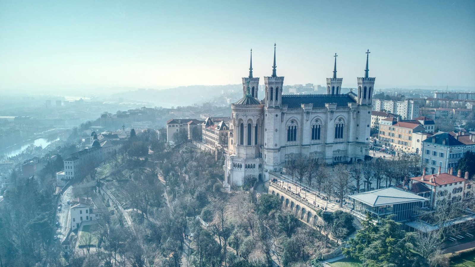 A pale stone basilica with four corner towers stands on a wooded hilltop above the city in morning haze