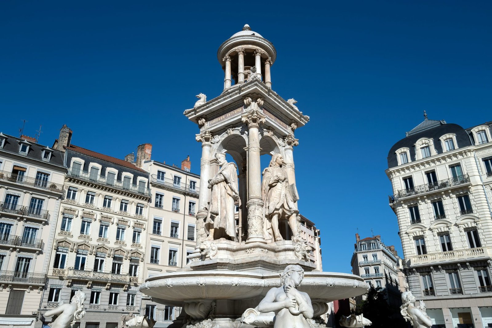 A white marble fountain with statues under a domed canopy, between cream apartment buildings and a deep blue sky
