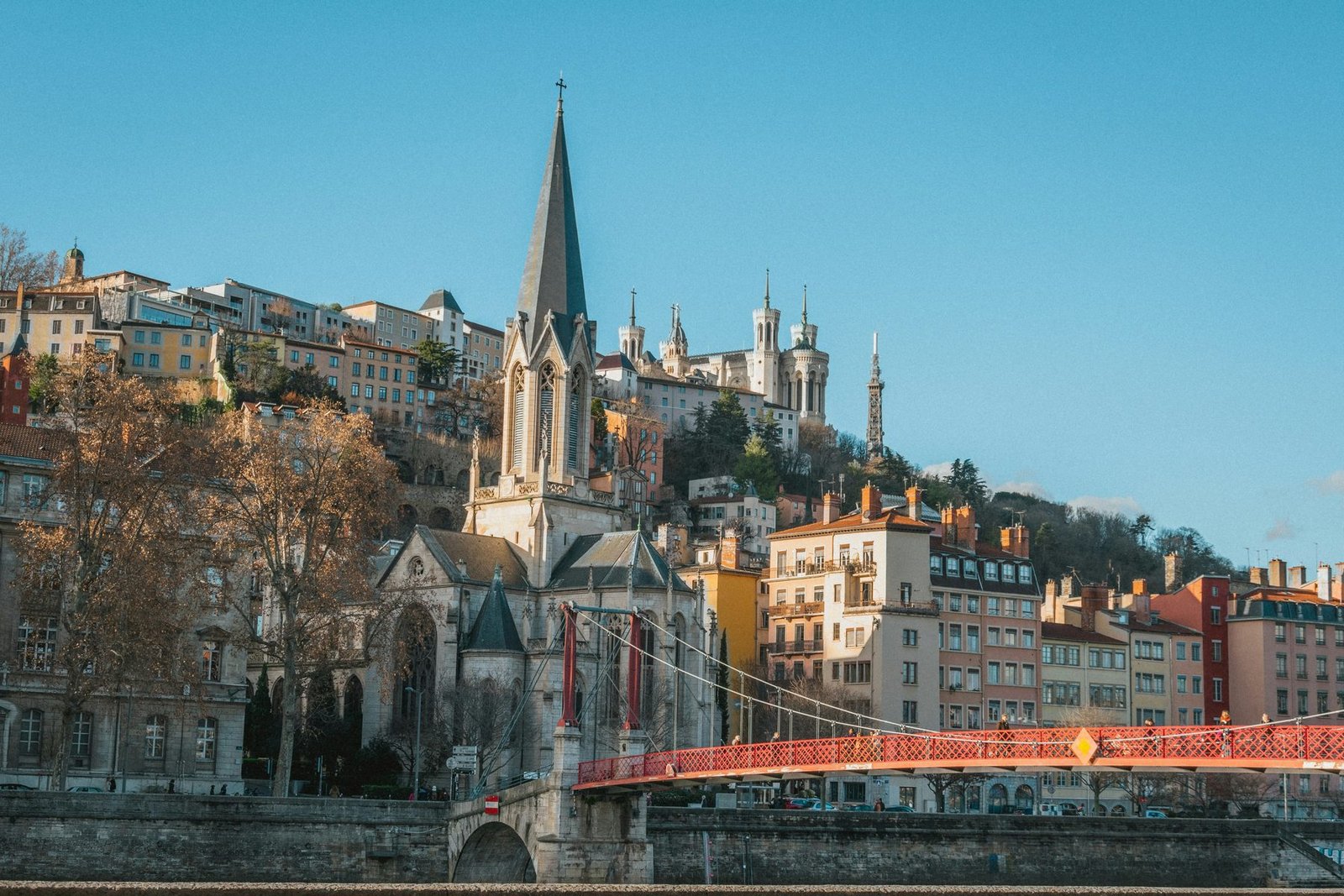 A church with a tall slate spire stands by a river and a red footbridge, with houses climbing a hill to a basilica behind