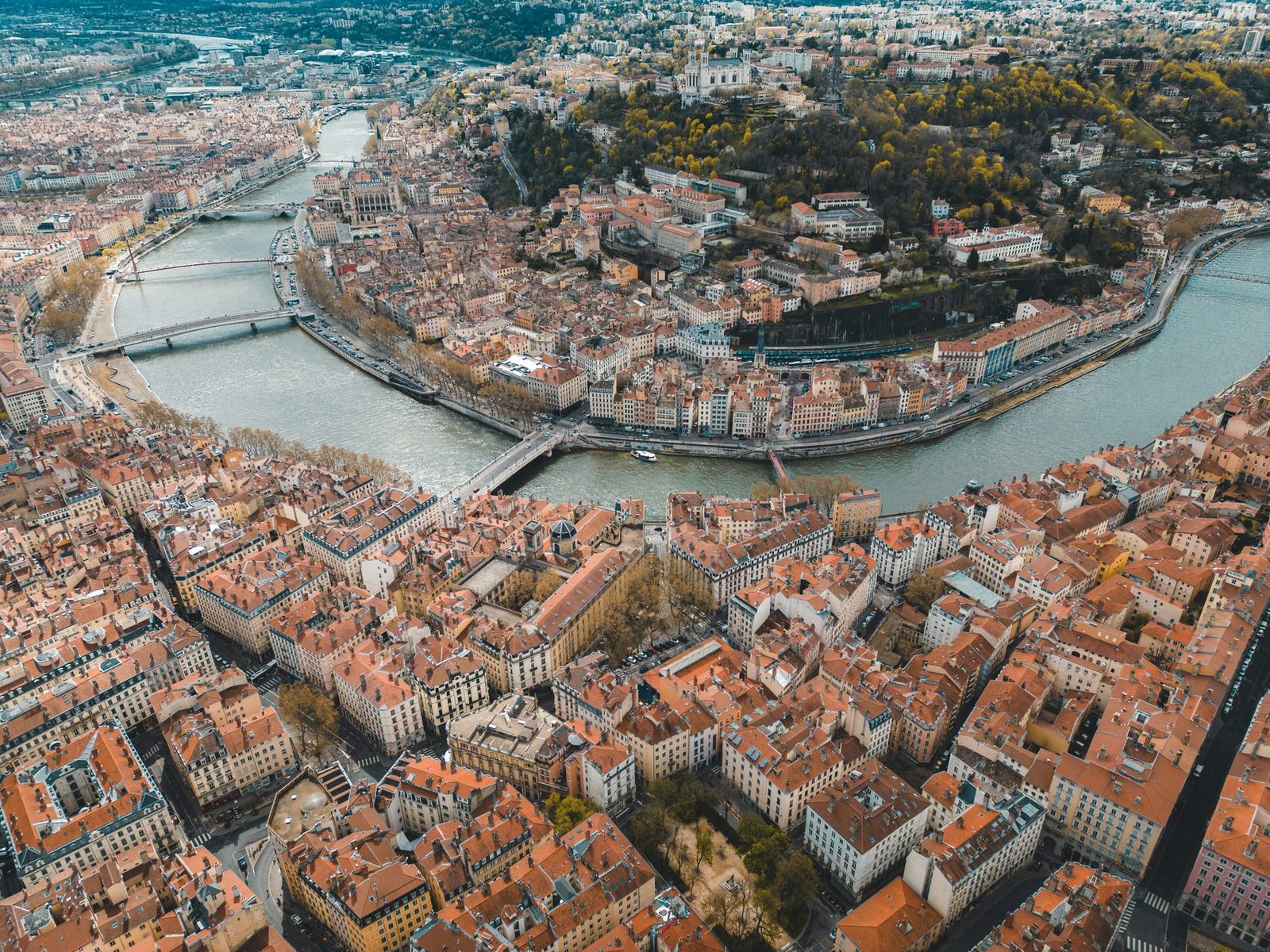 A river loops around a wooded hill through a dense city of red-tiled roofs, seen from high above