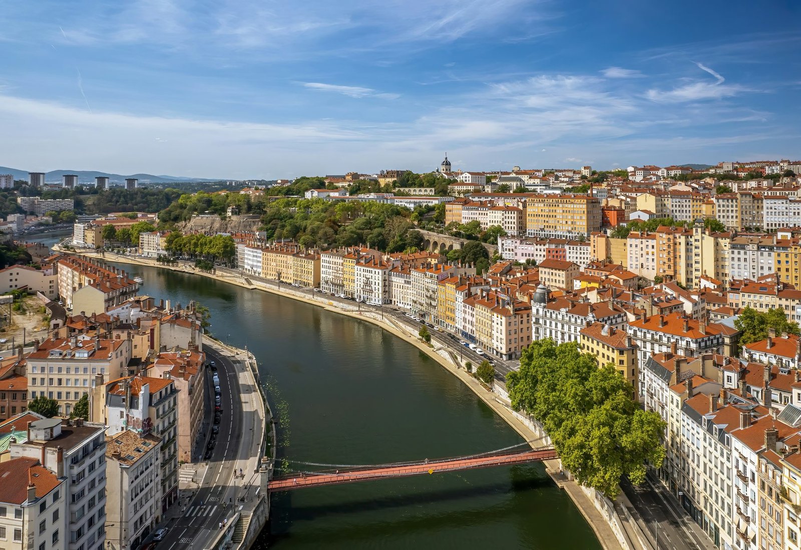 A green river bends between quays lined with pale and coloured houses with red roofs, crossed by a red footbridge