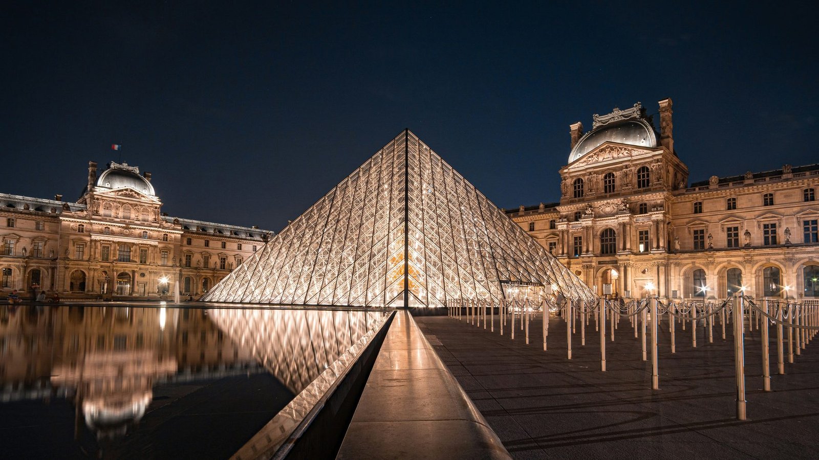 The glass pyramid and wings of the Louvre lit at night, reflected in the pool
