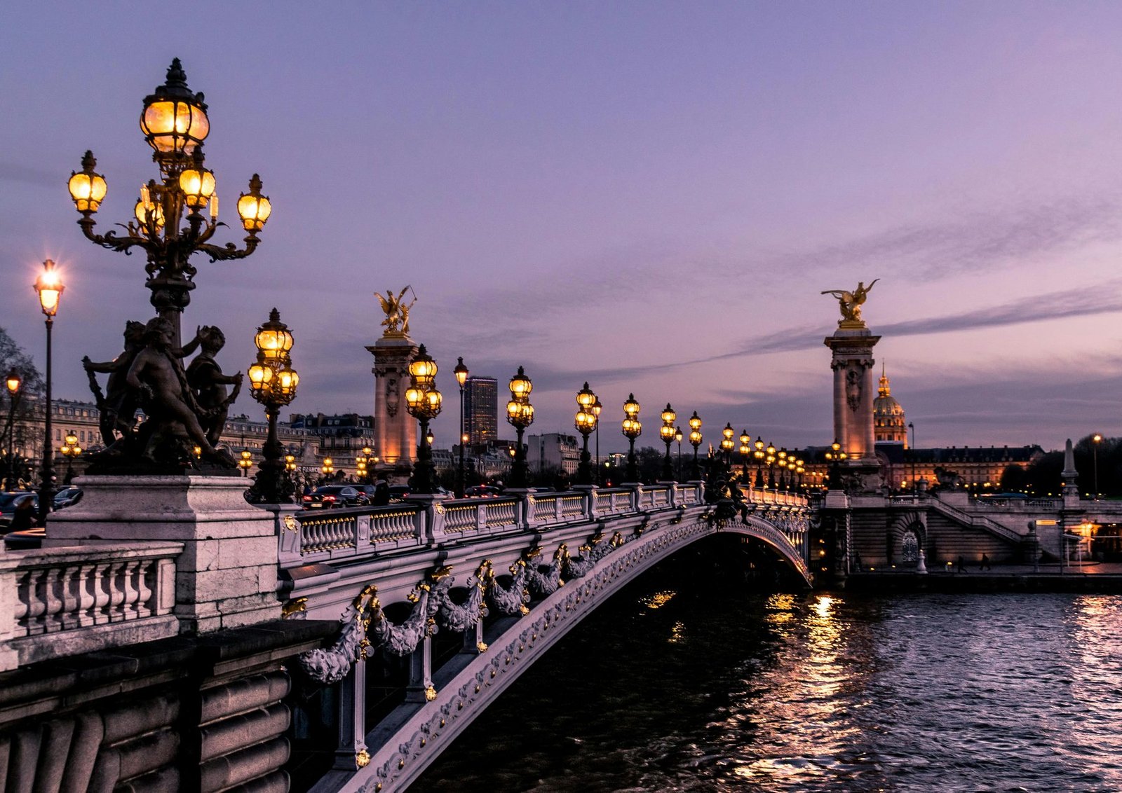 An ornate bridge with clusters of lit lamps and gilded statues on tall pillars crosses a river under a purple sky