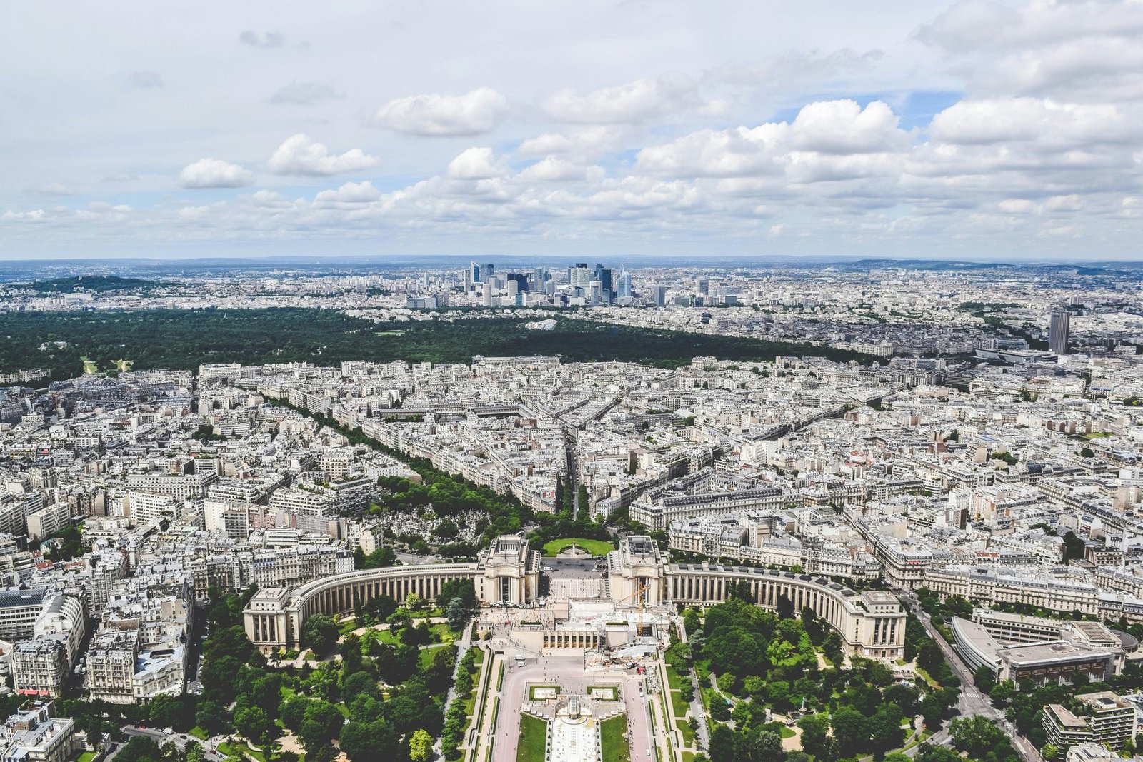 An aerial view of a curved colonnaded palace and its gardens, with dense city blocks, a large wooded park and distant towers beyond