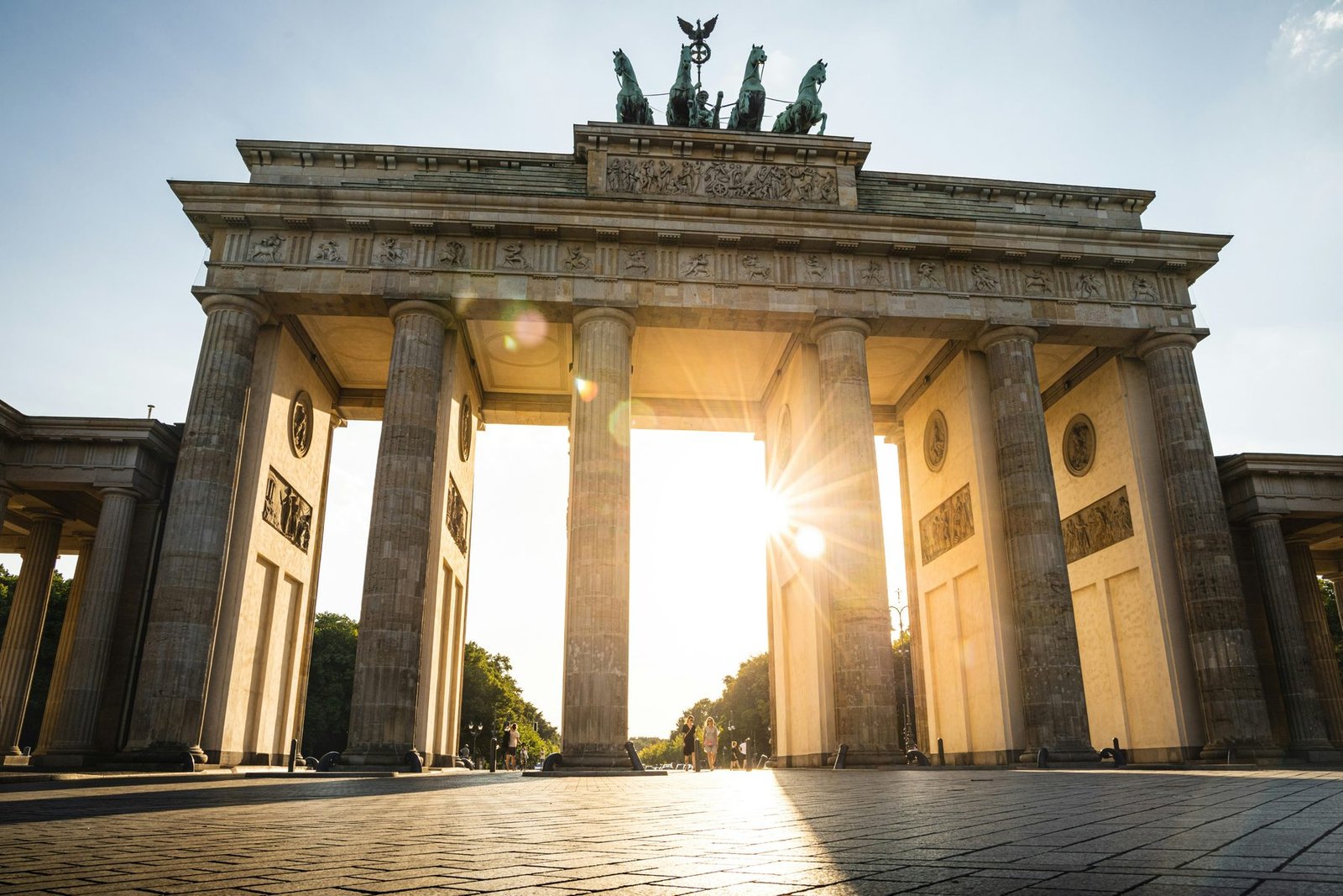 A neoclassical stone gate topped by a chariot sculpture, seen from a low angle with the sun shining between its columns