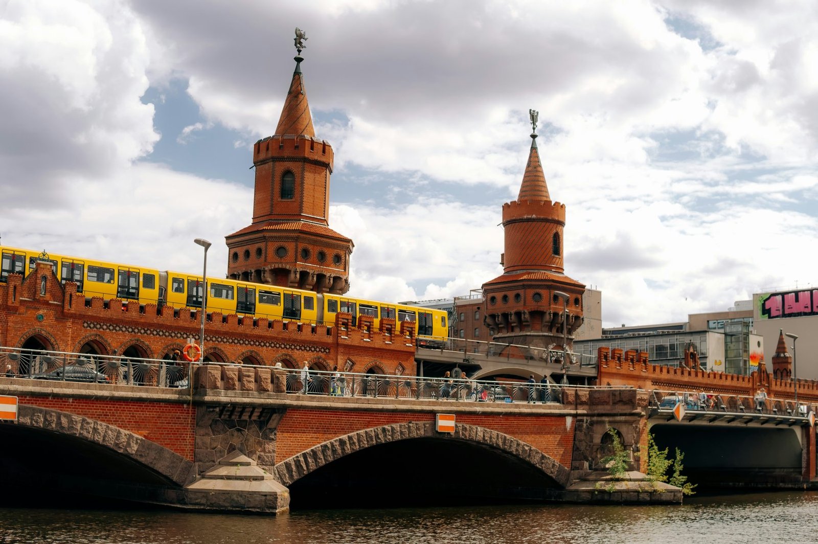 A red-brick double-deck bridge with two pointed towers spans a river while a yellow train runs along its upper level
