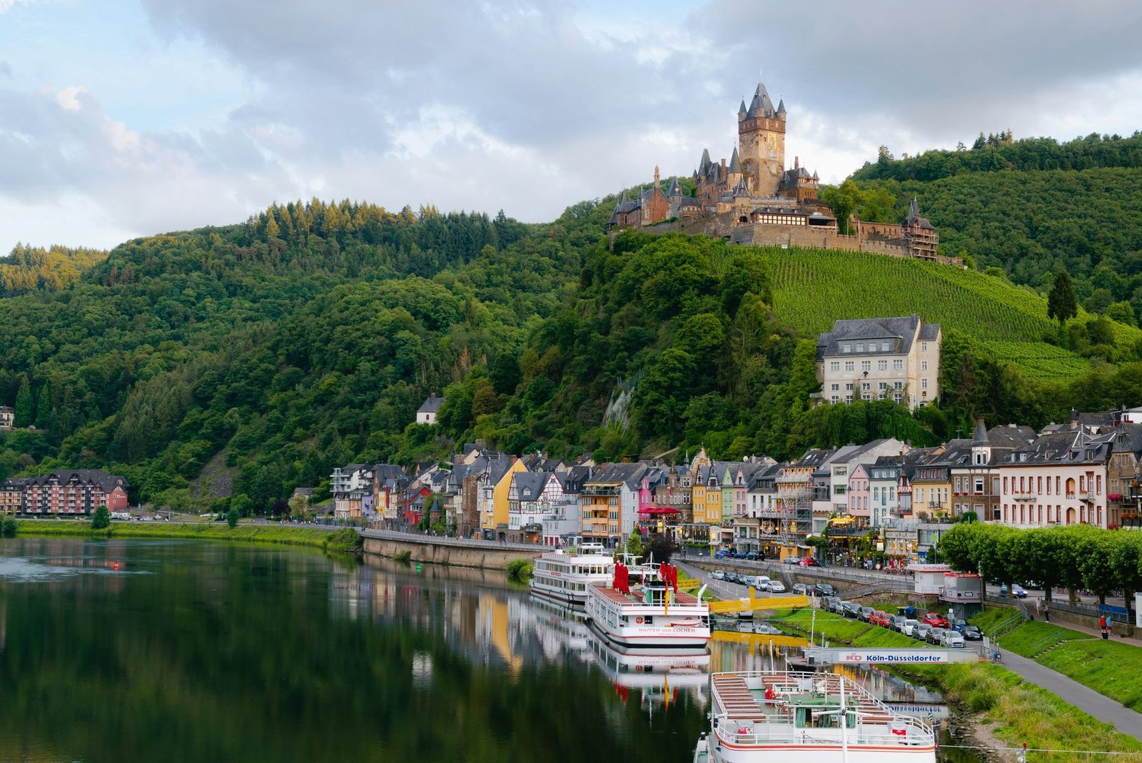 A turreted castle on a vine-covered hill above a river town with excursion boats moored along the bank