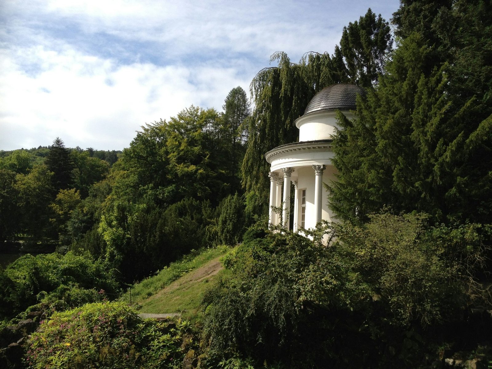 A small white rotunda with columns and a gray dome stands among dense trees on a slope