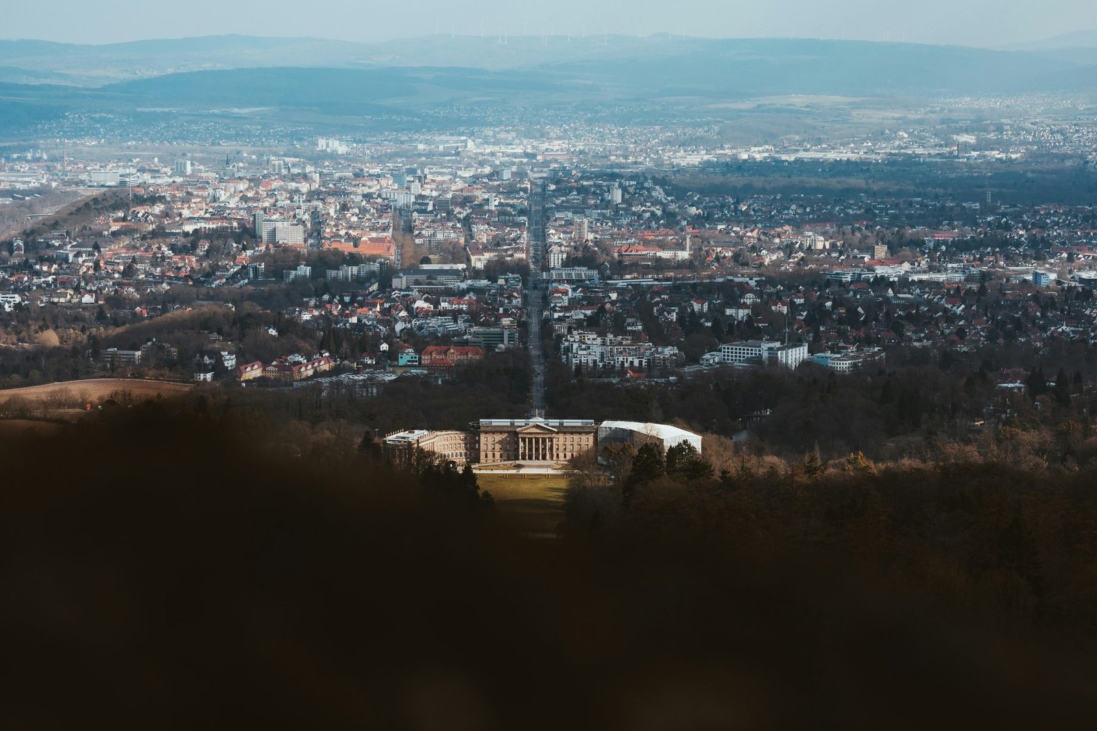 A palace at the foot of a wooded hill, with a straight avenue running from it into a city spread across the plain
