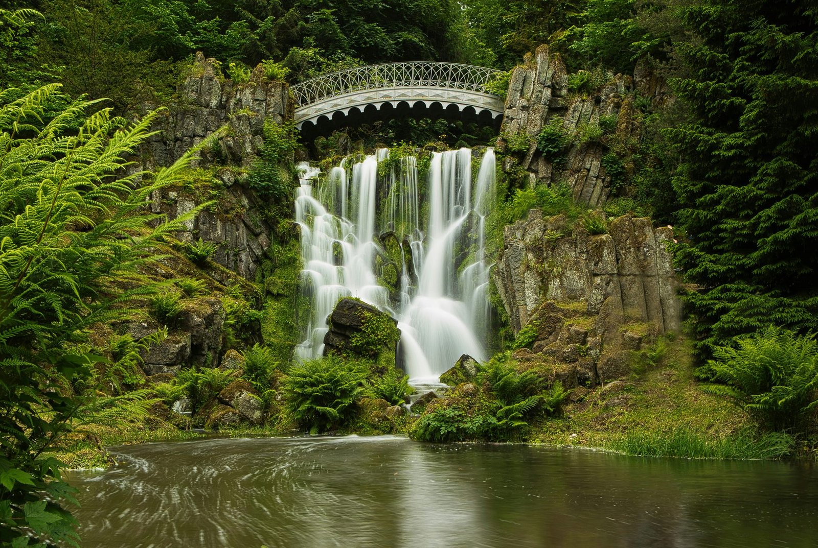 A waterfall drops over mossy rocks beneath a small arched iron bridge into a pool, framed by ferns
