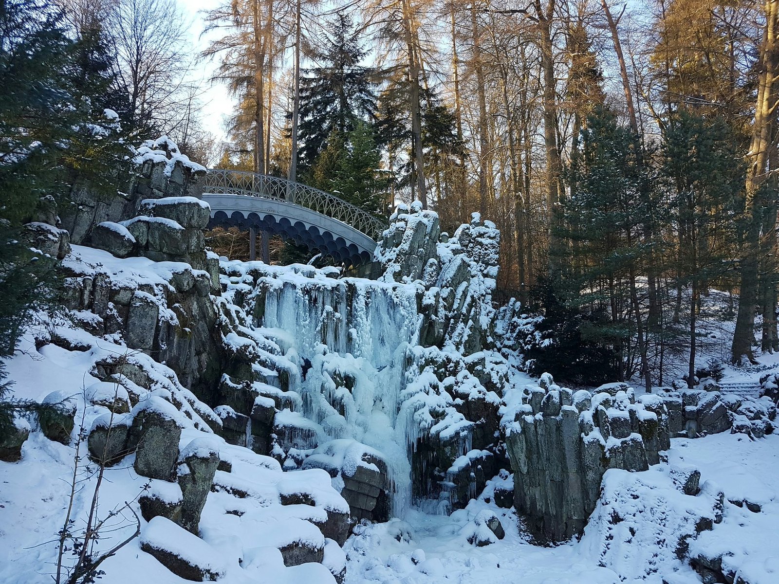 A frozen waterfall under a small arched bridge among snow-covered rocks and tall trees