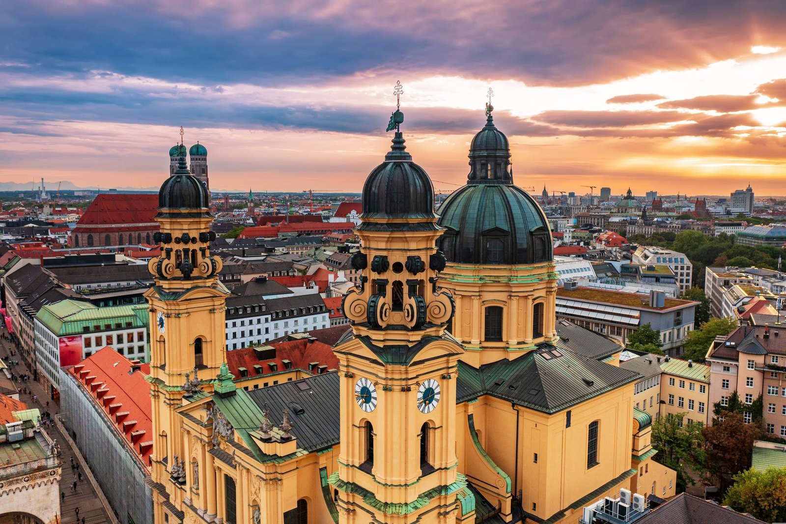 A yellow Baroque church with a dark green dome and twin towers seen from above, with city rooftops and an orange and purple sky