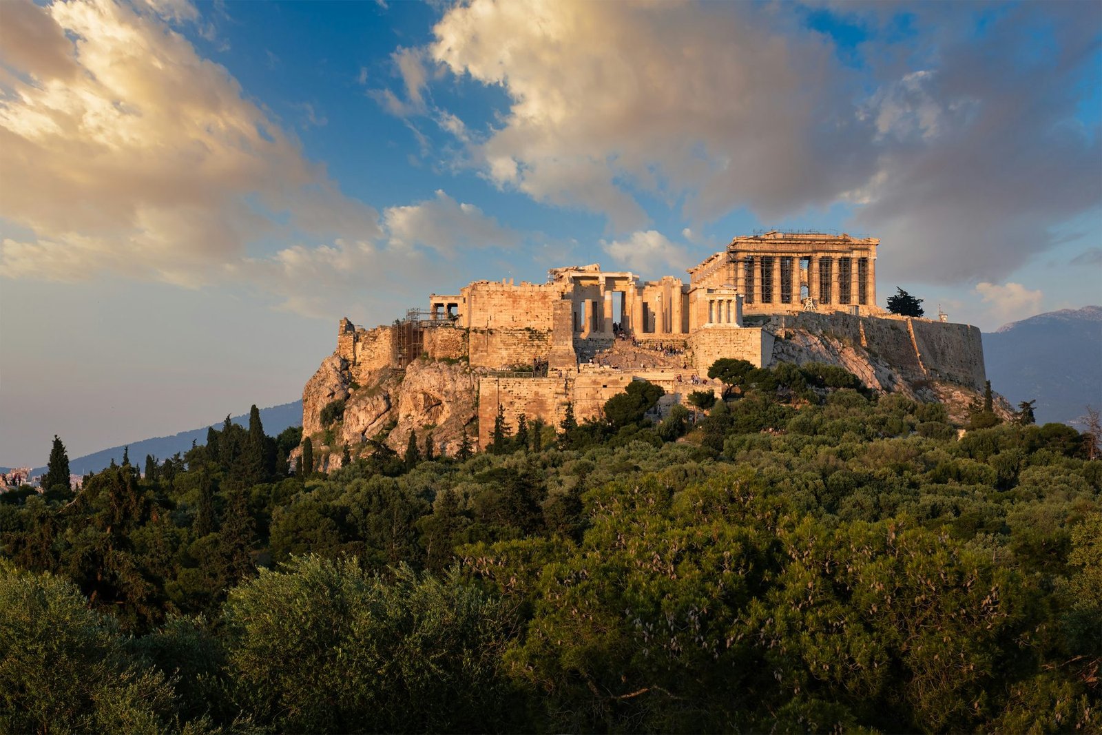 Temples and walls on a sheer rock glow orange above dark trees, under blue sky and large clouds