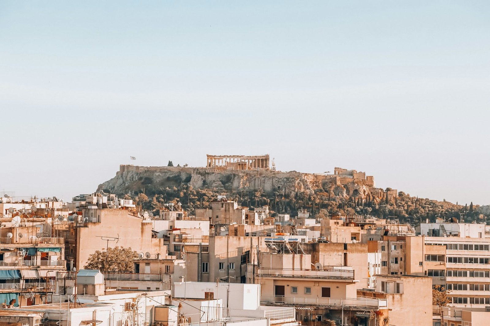 A flat-topped rocky hill with a columned temple rises over pale apartment blocks under a soft clear sky
