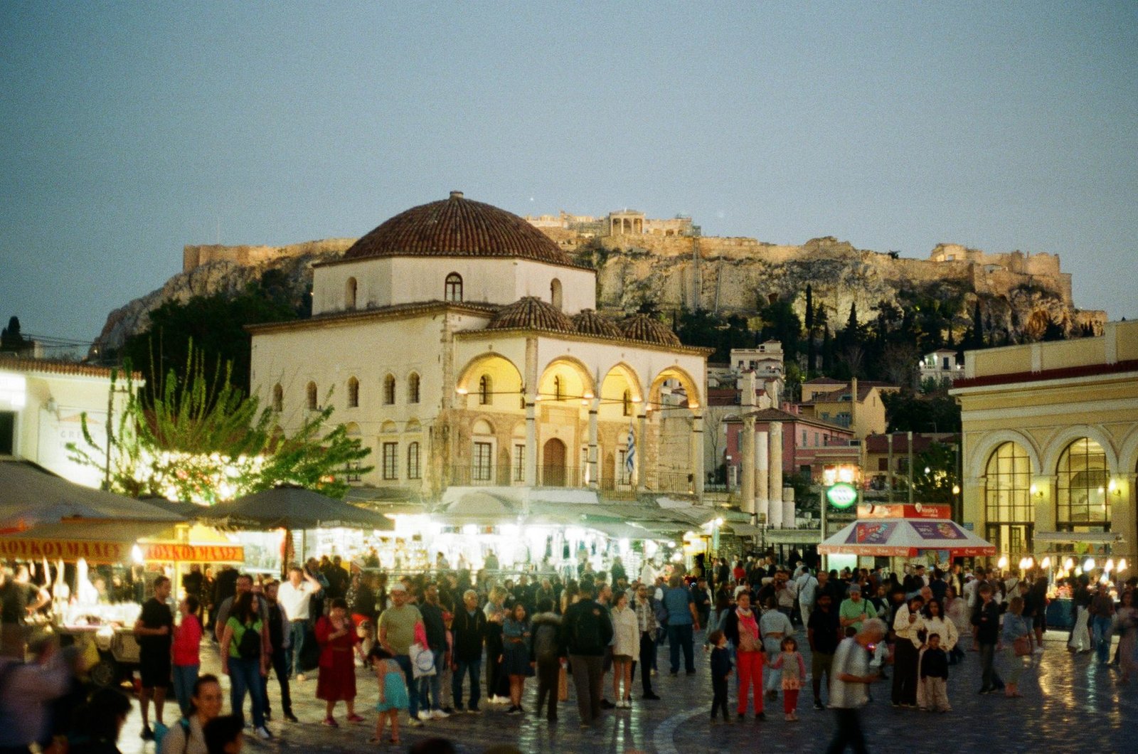 A crowd fills a square beneath lit market awnings and a domed building, with a floodlit rocky hill behind