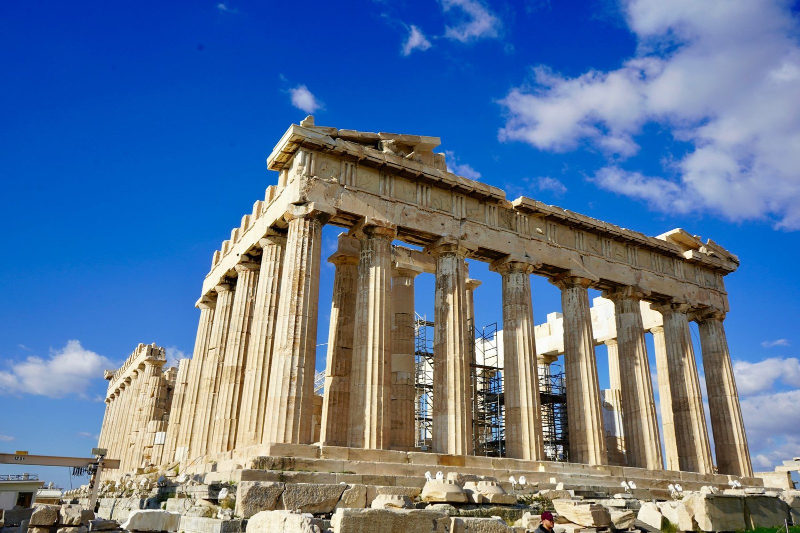 A marble temple with fluted columns is seen from one corner against deep blue sky and white clouds, with scaffolding inside