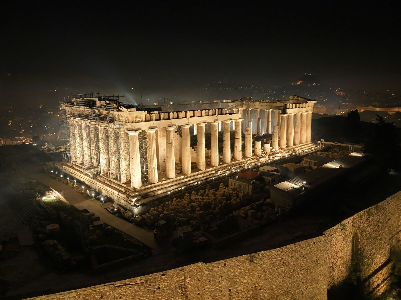 A lit marble temple with rows of columns glows on a dark hilltop with the city in darkness around it