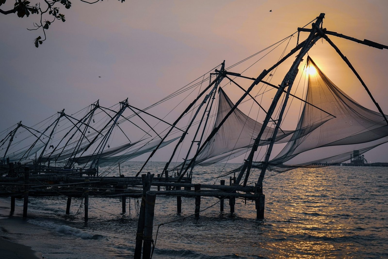 Large cantilevered fishing nets on wooden frames stand in silhouette over the water with the sun behind them