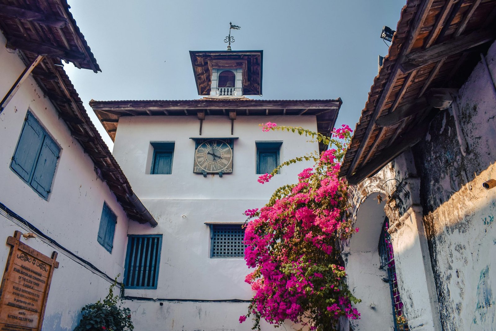 A whitewashed tower with a small belfry and weathervane rises between tiled-roof houses beside a flowering pink bougainvillea