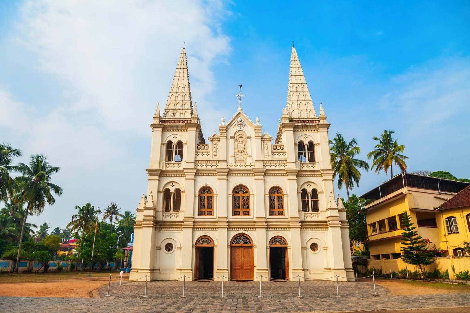 A cream-colored church with two tall spires and three arched doors stands among palms under a blue sky