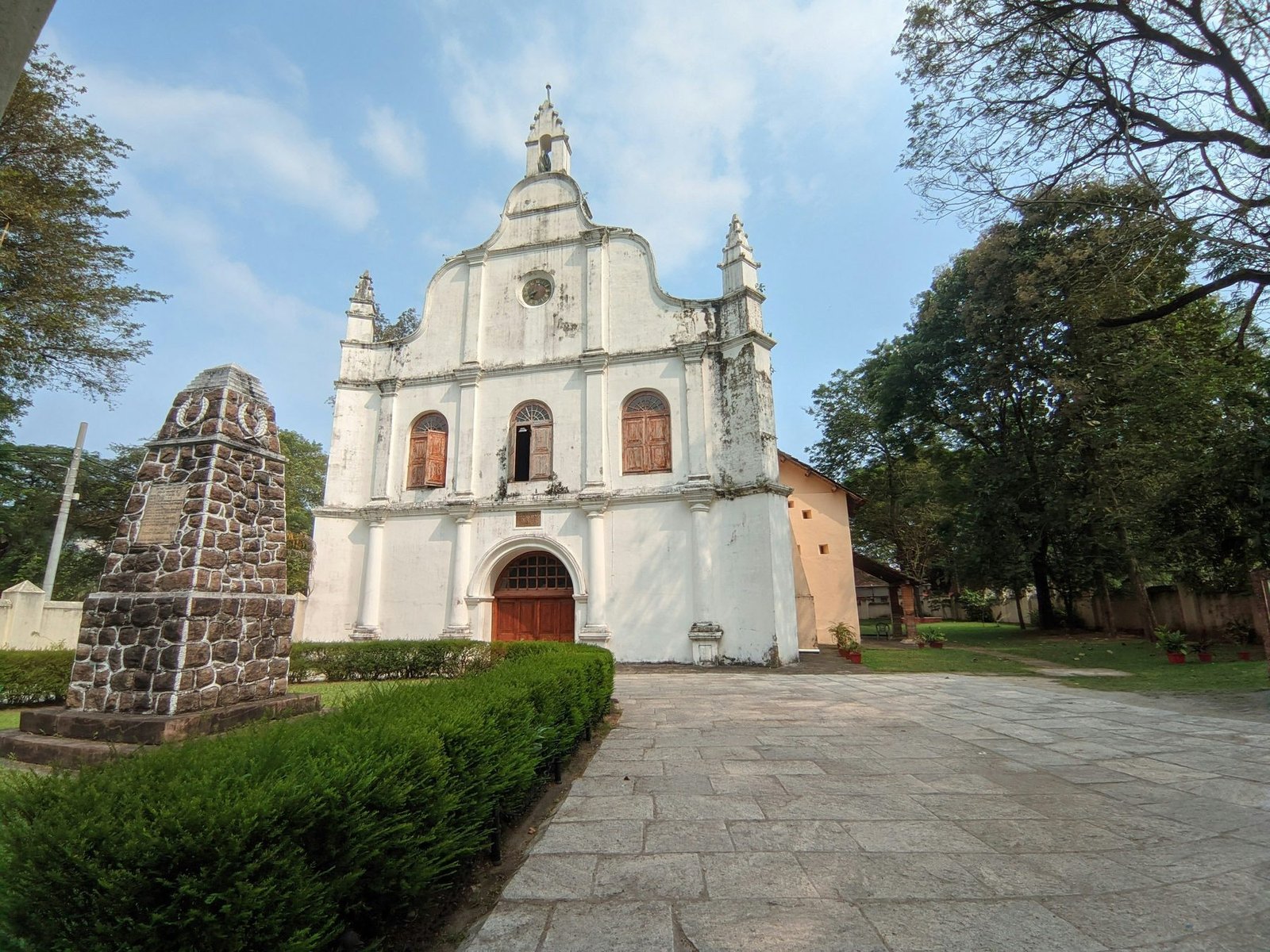 A plain white gabled church with a stepped front stands at the end of a stone path beside a small stone monument and a clipped hedge