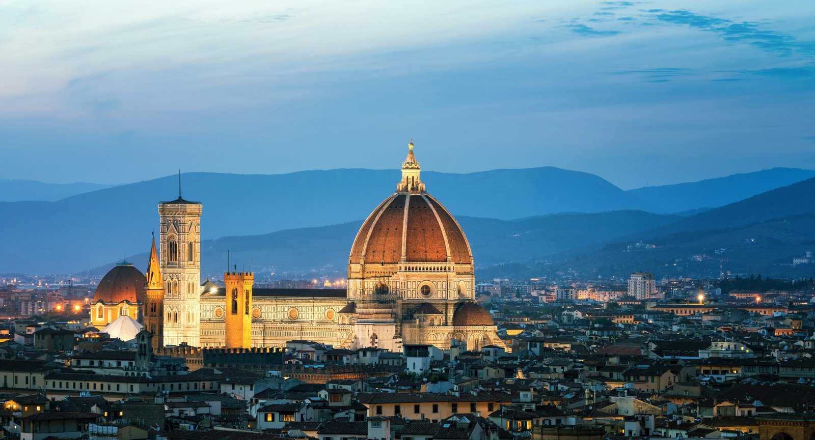 A cathedral with a large red-tiled dome and a tall bell tower lit above a dark city, with blue hills behind