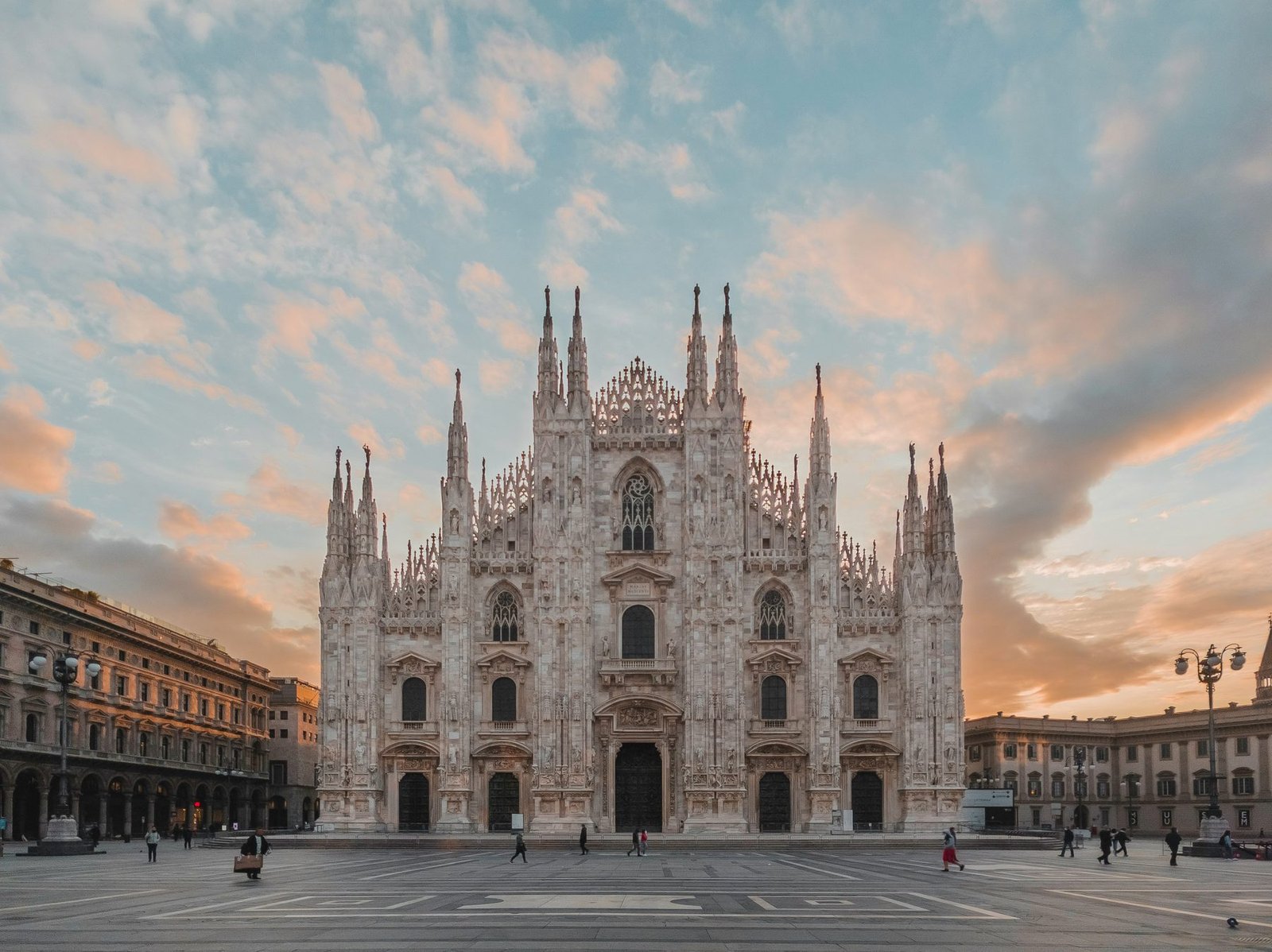 A white marble Gothic cathedral front with many spires facing a nearly empty square under a pink and blue sky
