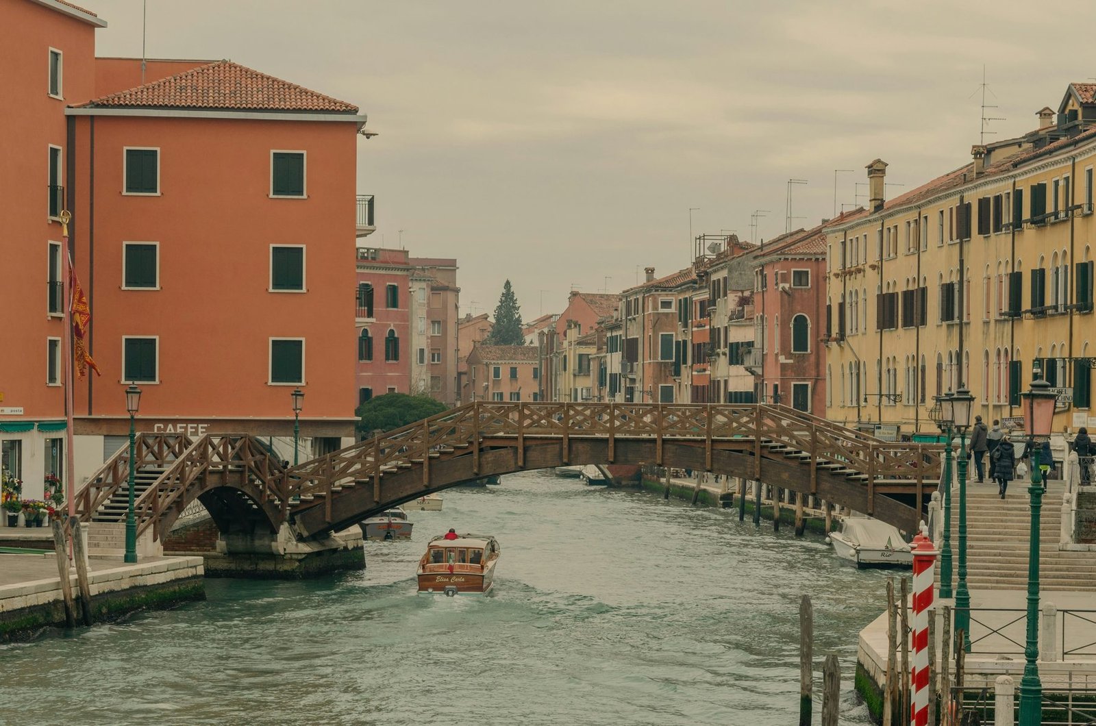 A wooden arched footbridge spans a canal between a terracotta-colored palace and a row of ocher buildings, with a motorboat passing below