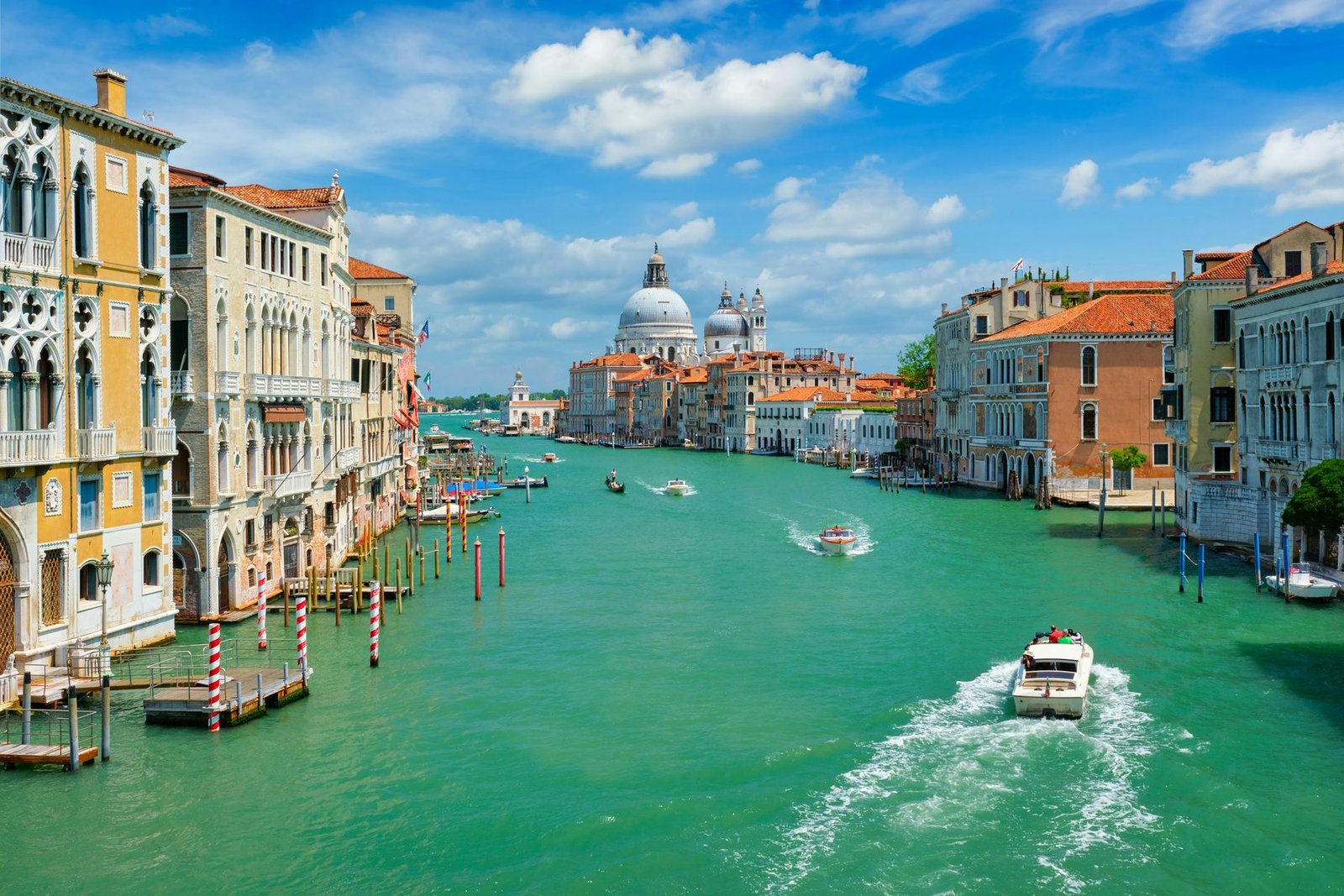 A motorboat leaves a white wake on green water between rows of palaces, with a domed church in the distance under a blue sky