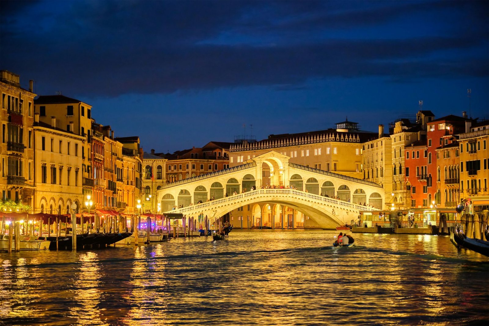 A covered stone arch bridge glows gold over a canal between lit palaces, with gondolas on the water under a dark blue sky