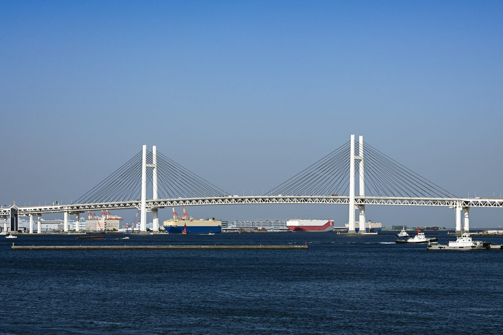 A long cable-stayed bridge with two white H-shaped towers crosses dark blue harbor water under a clear sky, with ships beneath