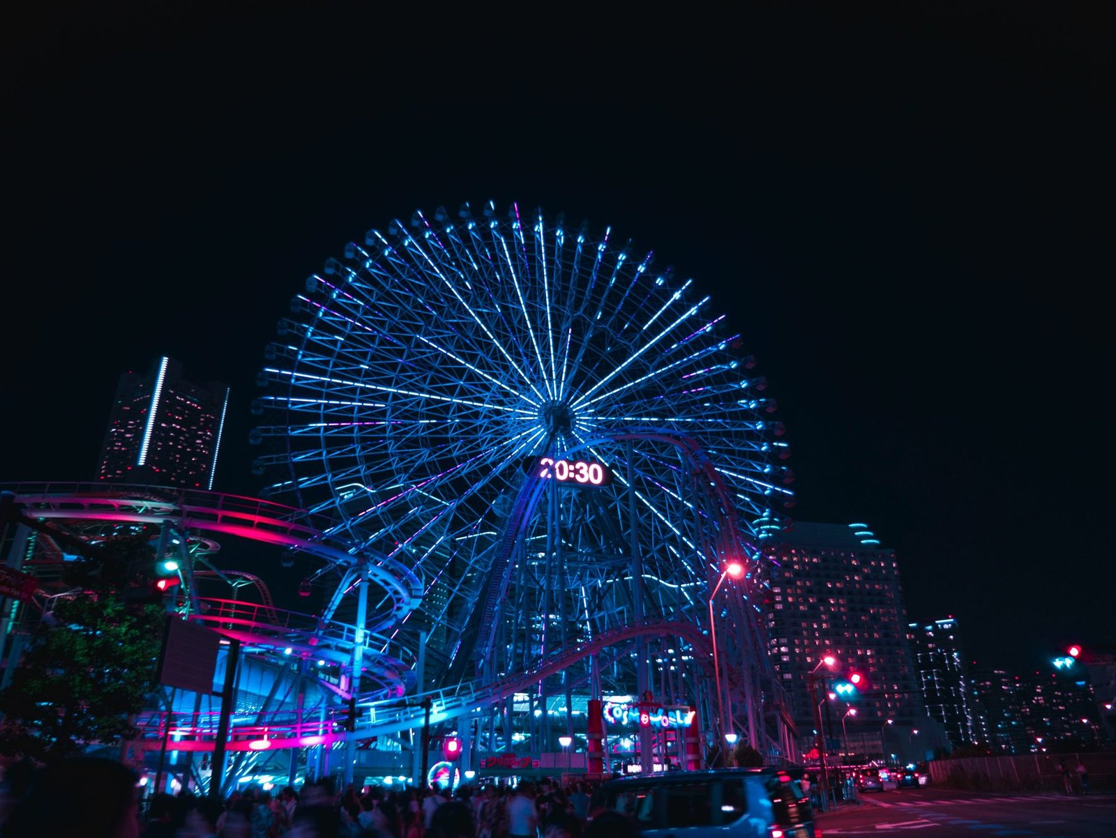 A giant observation wheel glows blue with a digital clock at its hub above pink and blue lit rides against a black sky