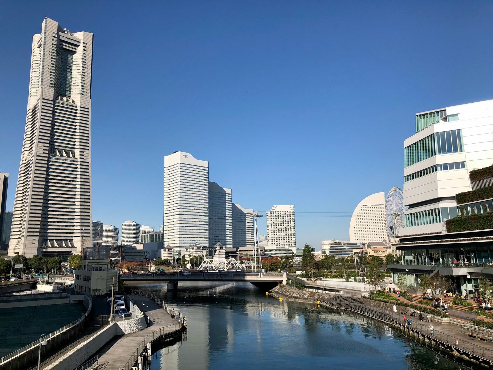 A tall tapering stone-gray skyscraper rises at left above a canal with a promenade, white towers and a bridge under a deep blue sky