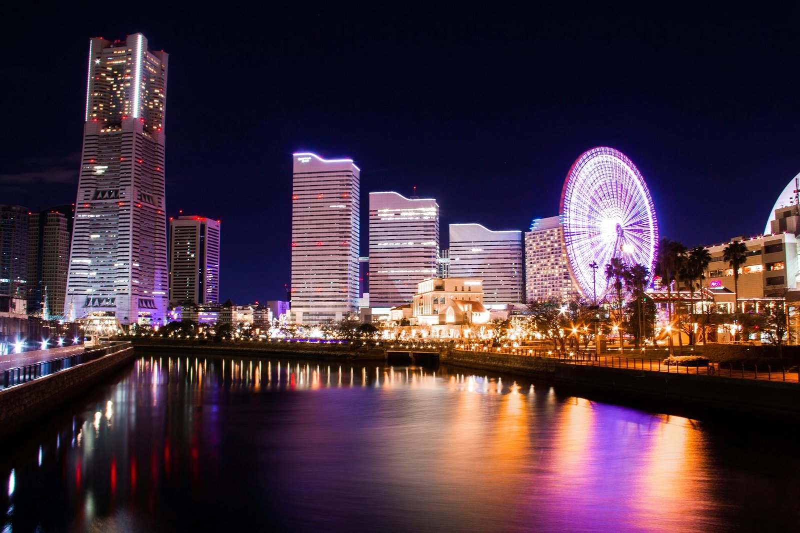 A tall tower, stepped high-rises and a purple-lit observation wheel are reflected in a canal under a dark sky
