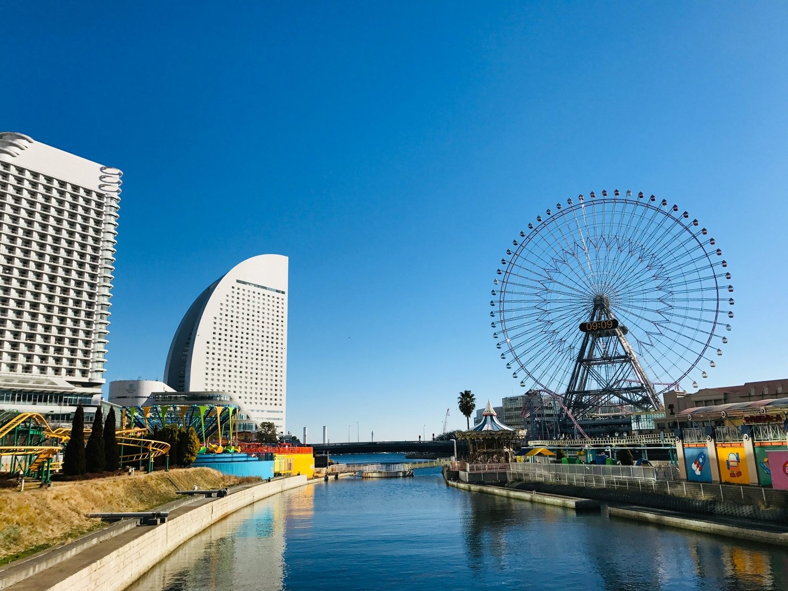 A large Ferris wheel and a sail-shaped white hotel tower stand beside a canal under a clear blue sky